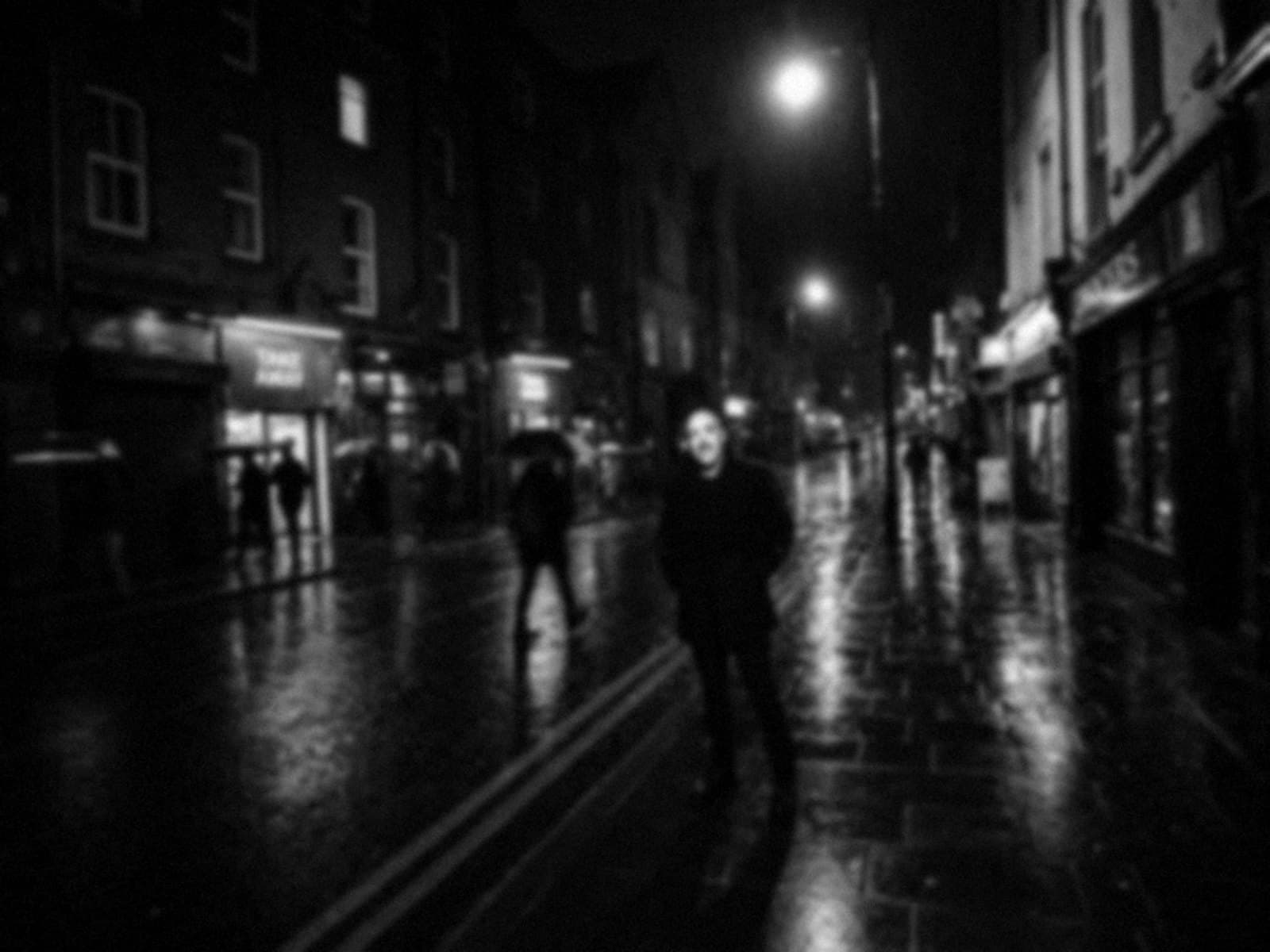 Night photograph of Jonny Campbell standing still on a rainy Belfast street while passers-by blur past