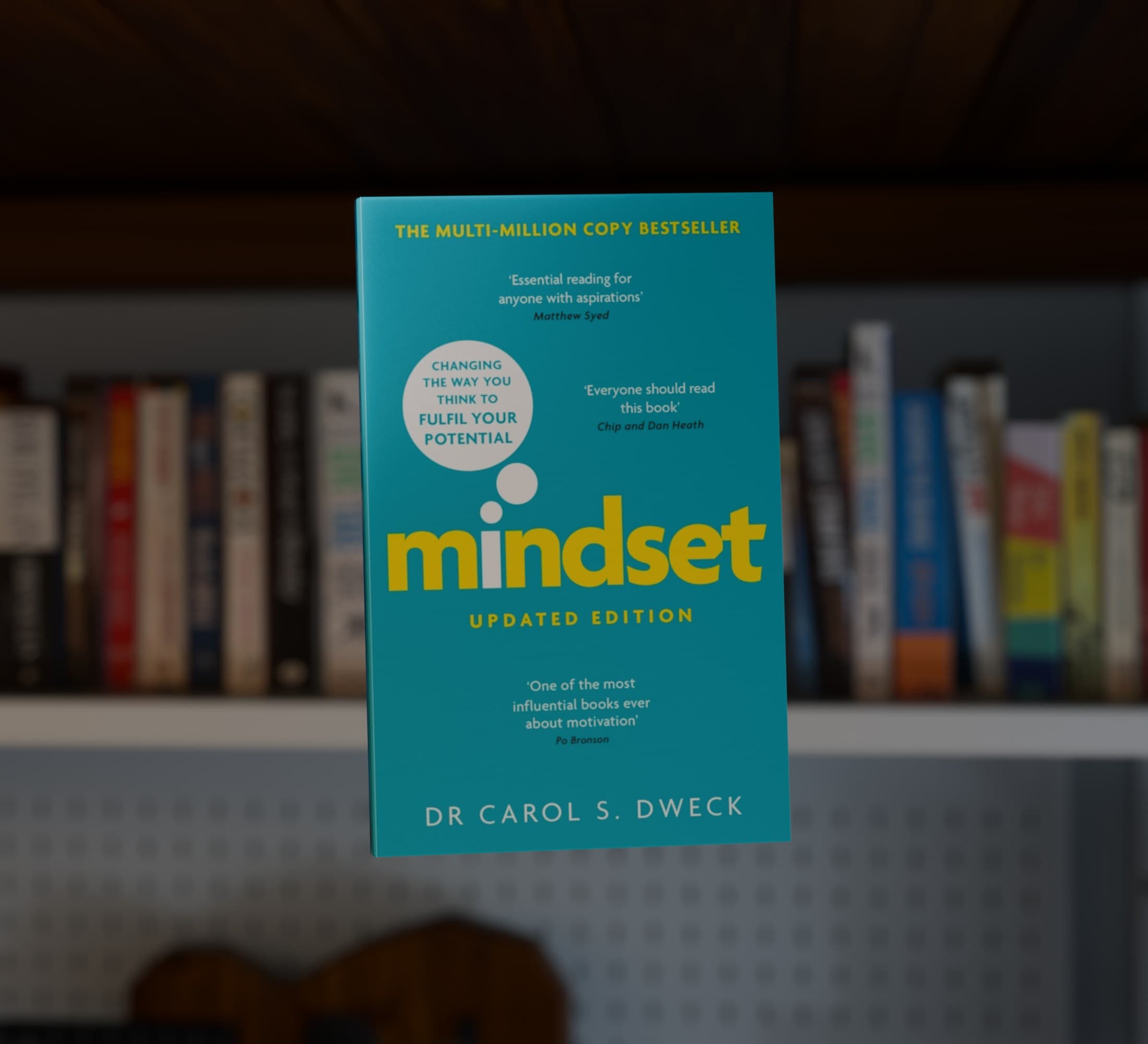 A render of a teal copy of Mindset by Carol S. Dweck, floating face on in front of a shelf of books, with the shelf and a white pegboard behind it out of focus.