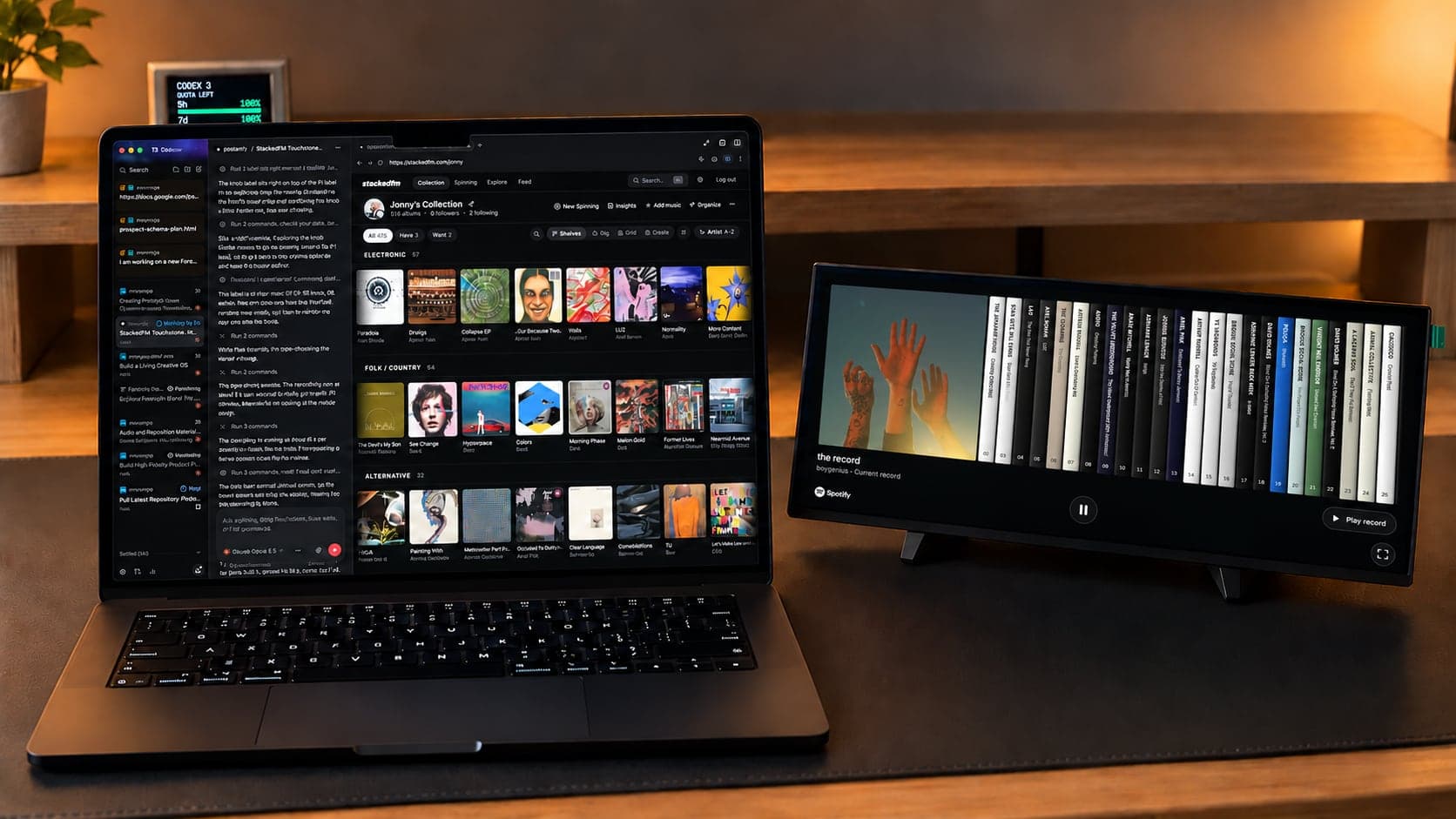 A laptop and a wide touchscreen on a small stand, side by side on a desk. The laptop shows StackedFM's collection beside a chat with a coding agent, and the touchscreen shows StackedFM's Spines view.