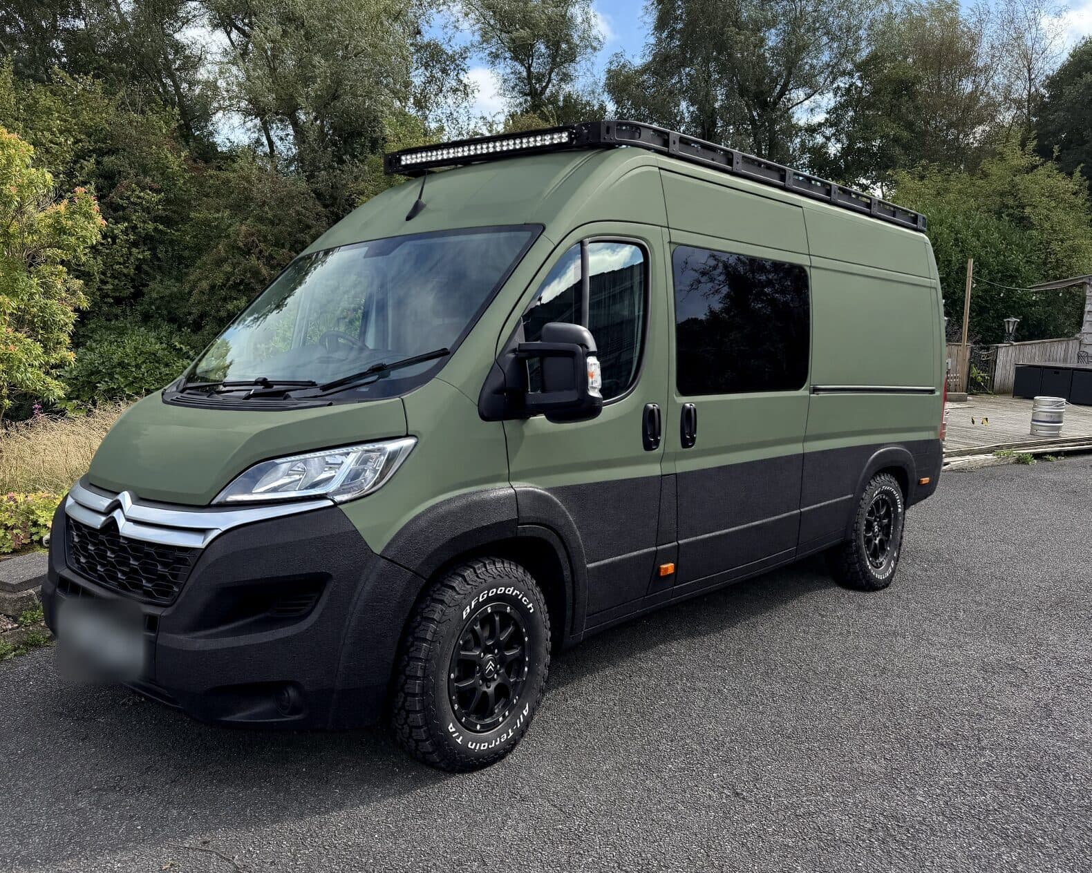 AI mock-up of the same van in the same spot, in satin military green with black lower panels, a tinted window in the sliding door, a roof rack with a light bar, black wheels and all-terrain tyres.